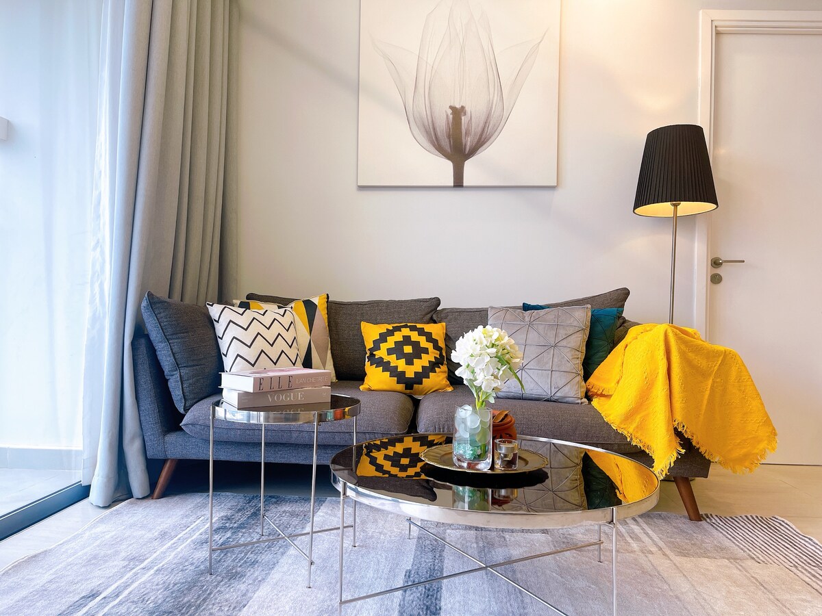 A modern living area is highlighted by a large floral artwork above a comfortable gray sofa adorned with decorative cushions in various patterns and colors. A sleek coffee table sits in front of the sofa, accompanied by a vase with fresh flowers. Natural light enters through a window, creating a bright ambiance.