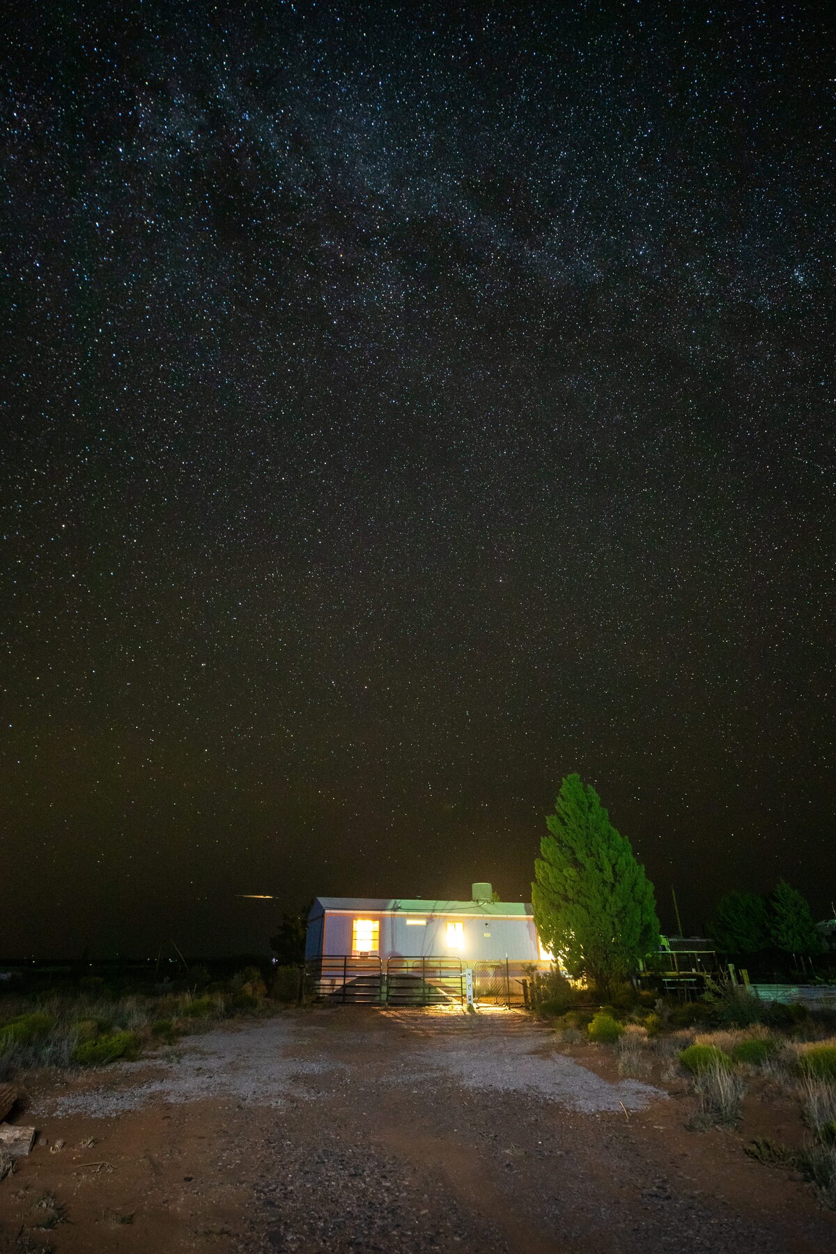 A serene night scene is captured, showcasing a house illuminated by warm light, beneath a vast sky filled with countless stars. The surrounding landscape features sparse vegetation and a gravel path leading to the entrance, enhancing the sense of tranquility in the remote setting.