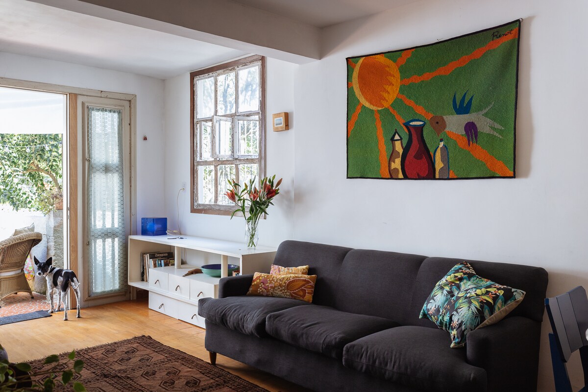 A sunny living room is highlighted by a large window and a door leading to the outside. A dark sofa with decorative cushions faces a wooden floor and a vibrant wall tapestry. A small shelf displays books, while fresh flowers are arranged on a nearby table.