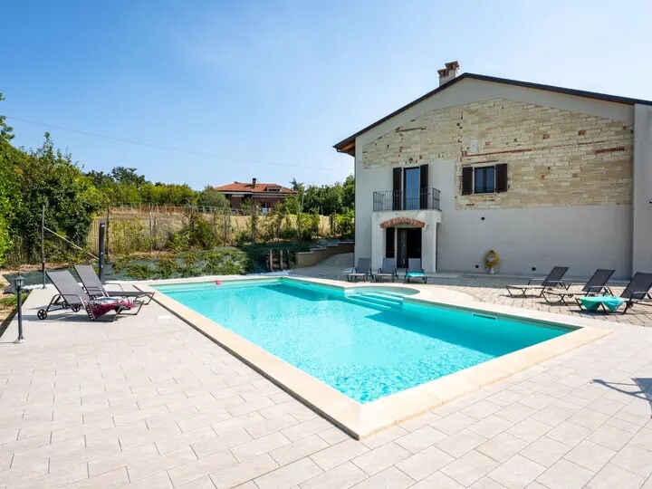 An outdoor swimming pool is surrounded by a spacious deck, featuring several lounge chairs for relaxation. The villa, with textured stone walls, stands in the background, complemented by a tranquil landscape of greenery. Sunlight bathes the area, creating a welcoming atmosphere.