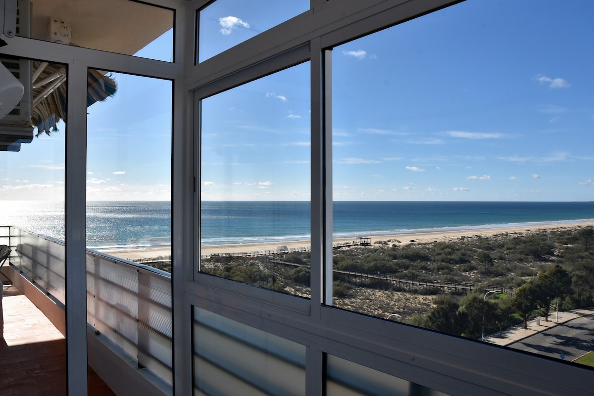 A panoramic view showcases a serene coastline, with soft sandy beaches meeting gentle ocean waves. Large glass windows provide ample natural light and an unobstructed view of the clear blue sky above and the tranquil sea below.