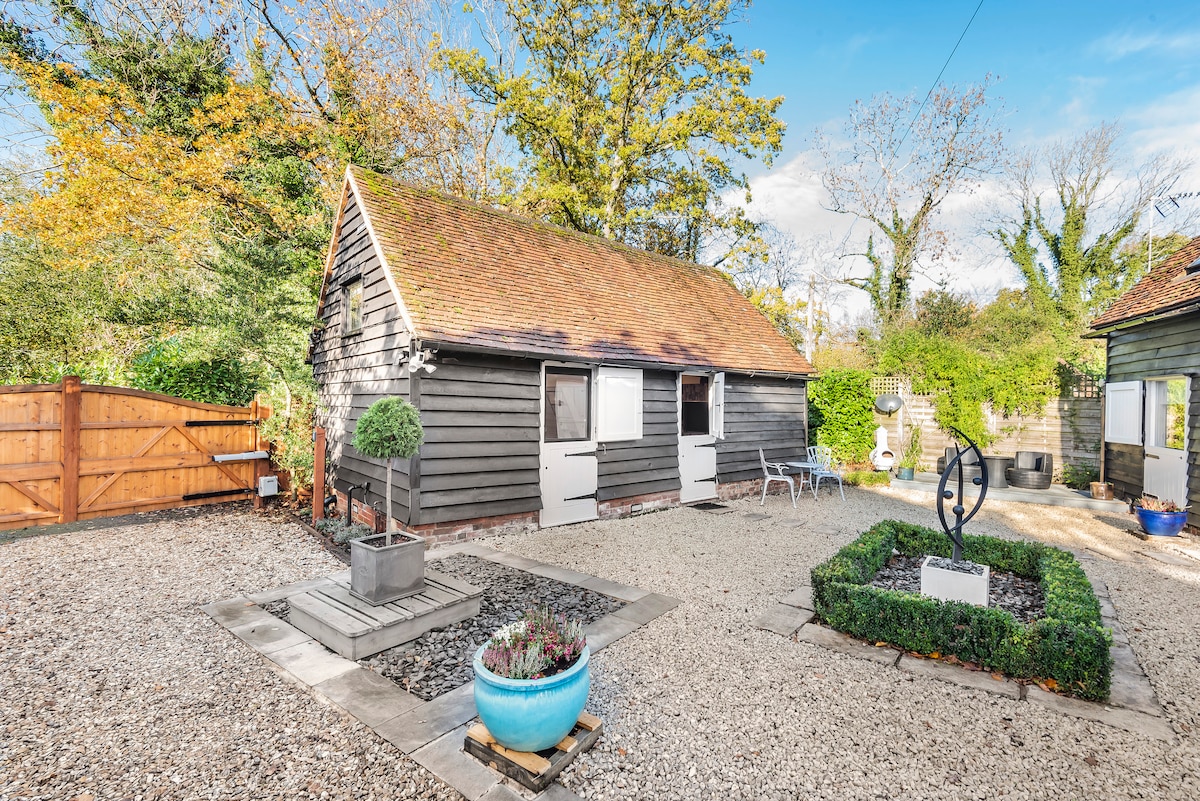 A charming converted barn exterior showcases a gravel courtyard surrounded by lush greenery. Flanking the entrance are two large windows framed by white accents. A landscaped area features a circular design with planters, complemented by a sculpture as a focal point amidst the serene setting.