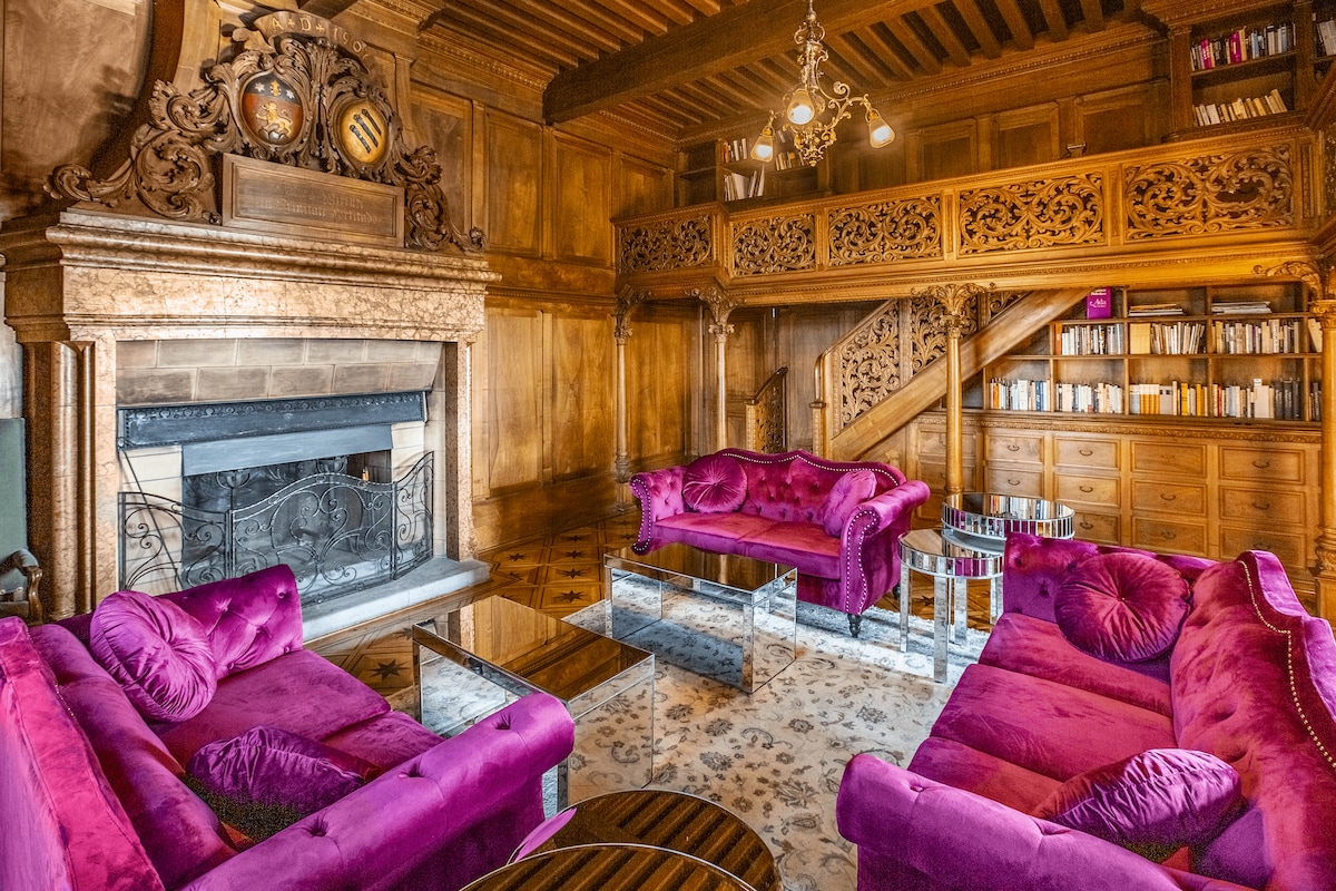 A grand lounge area features rich wooden paneling and intricate detailing. Two plush purple sofas are arranged around a glass-topped coffee table on a decorative rug, with a marble fireplace adding a classic touch. A staircase leads to a lofted space lined with bookshelves.