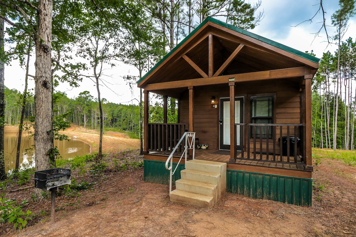 The exterior of a rustic cabin features a covered porch with steps leading down to the ground. To the side, a charcoal grill is available. Surrounding trees and a calm pond offer a natural setting, enhancing the sense of seclusion.