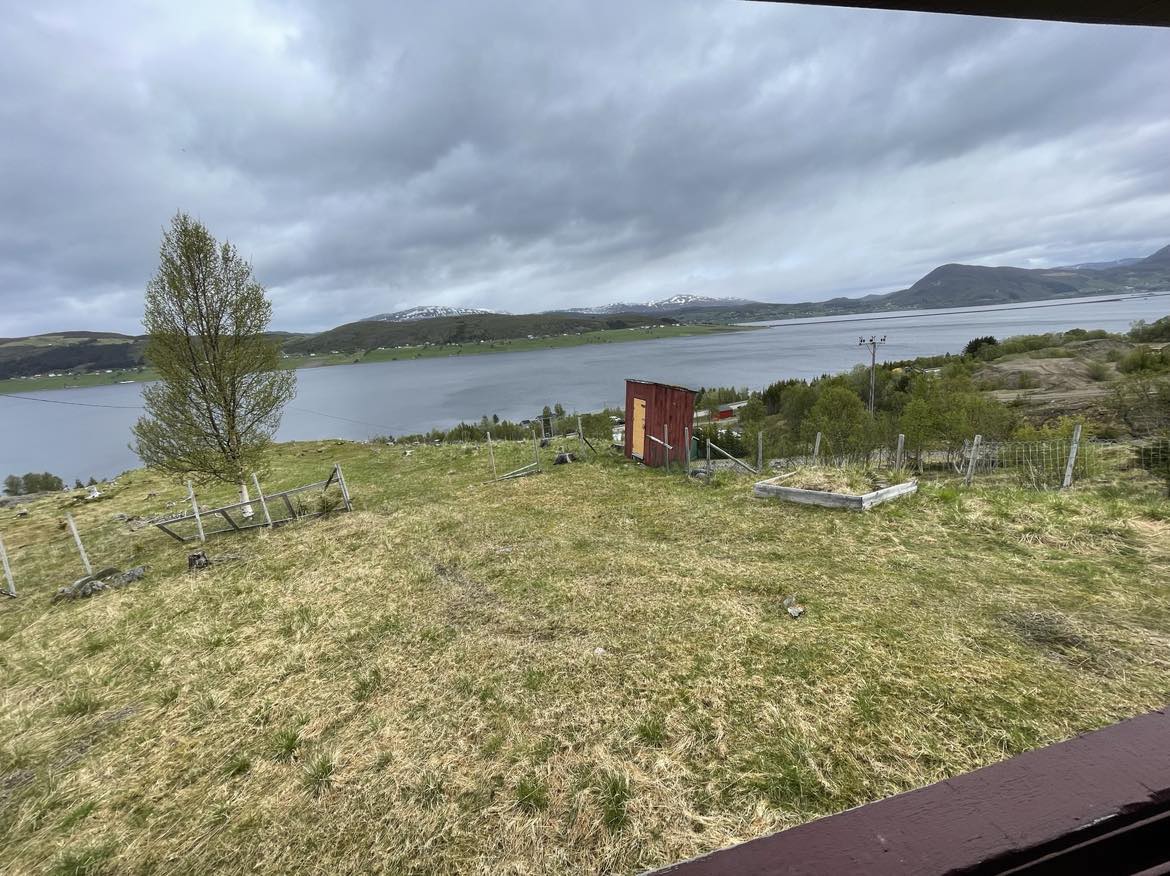 A panoramic view captures a tranquil landscape with a body of water stretching into the distance, bordered by rolling hills and snow-capped mountains. A small red shed is visible on the land, surrounded by green grass and scattered fencing.