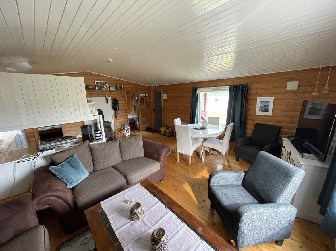 An open living area features a combination of brown and gray furniture, including a spacious sectional sofa and two armchairs. A dining table with white chairs is positioned near a window. Natural wood paneling and light-colored ceilings enhance the cozy ambiance.