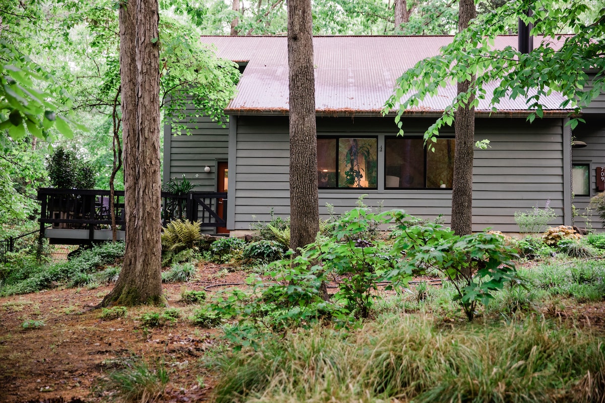 A contemporary wooden home is nestled among tall trees, surrounded by lush greenery and natural landscaping. Large windows reflect the forest atmosphere, while a small deck area is visible in the background, offering an inviting connection to the outdoors.