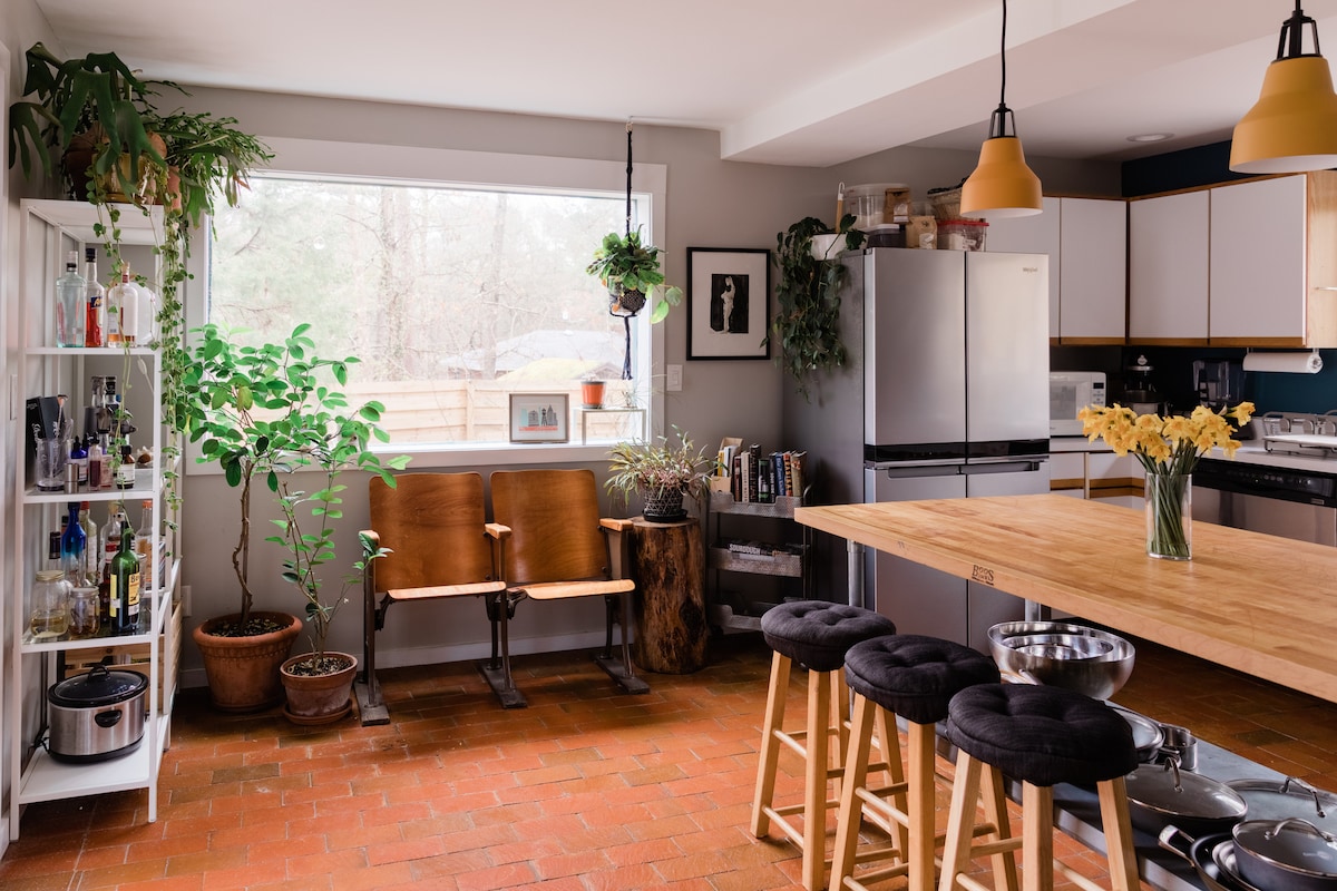 The kitchen area features natural light flowing through a large window, illuminating a wooden island with bar stools. Potted plants, artwork, and shelving with various items create a welcoming atmosphere, while modern appliances are positioned in a functional layout.