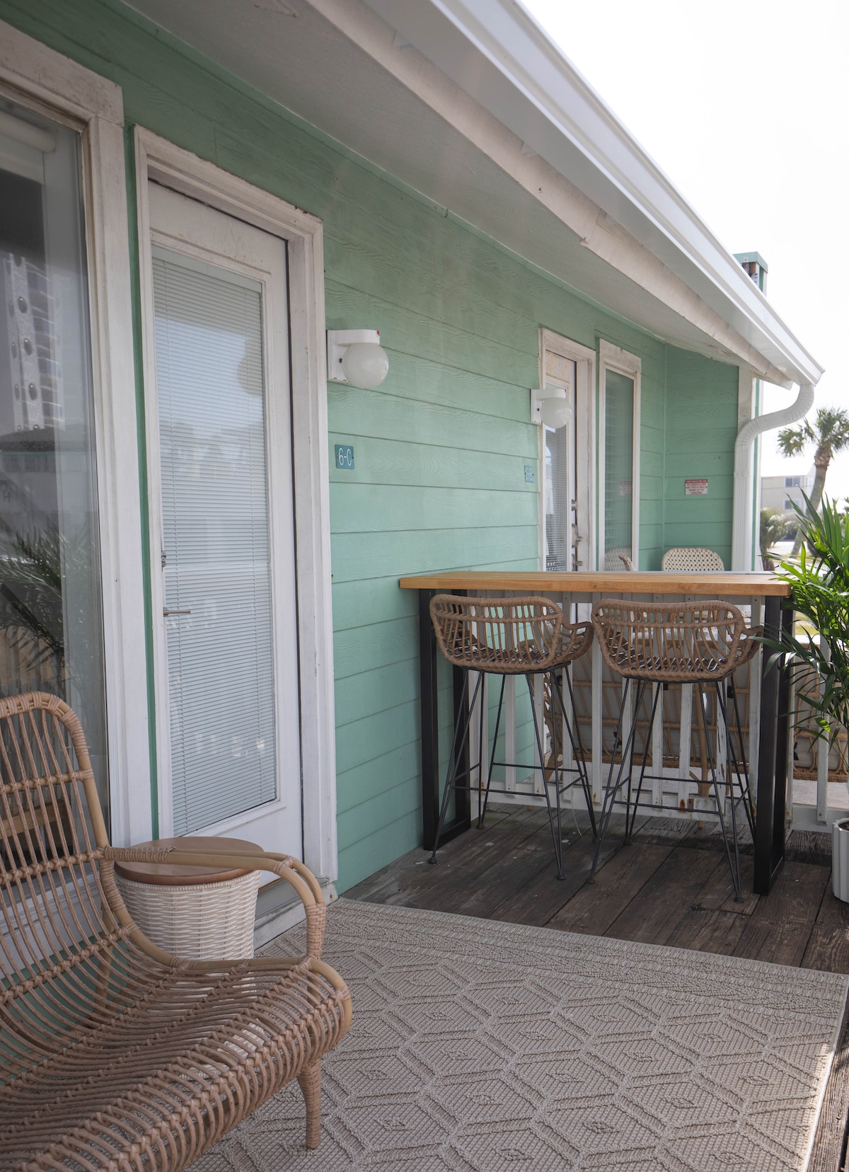 A cozy outdoor space features a light green exterior and a small bar table with four woven bar stools. A comfortable chair is positioned nearby, with a textured rug visible on the wooden deck. Lush plants are placed around the area, enhancing the inviting atmosphere.