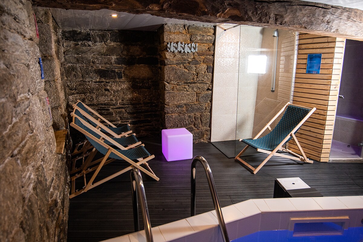 A relaxing spa area features stone walls and wooden accents. Two lounge chairs are positioned beside a heated pool, which exudes a calming ambiance. Soft lighting is provided by an illuminated cube, enhancing the serene atmosphere near the sauna and steam room.