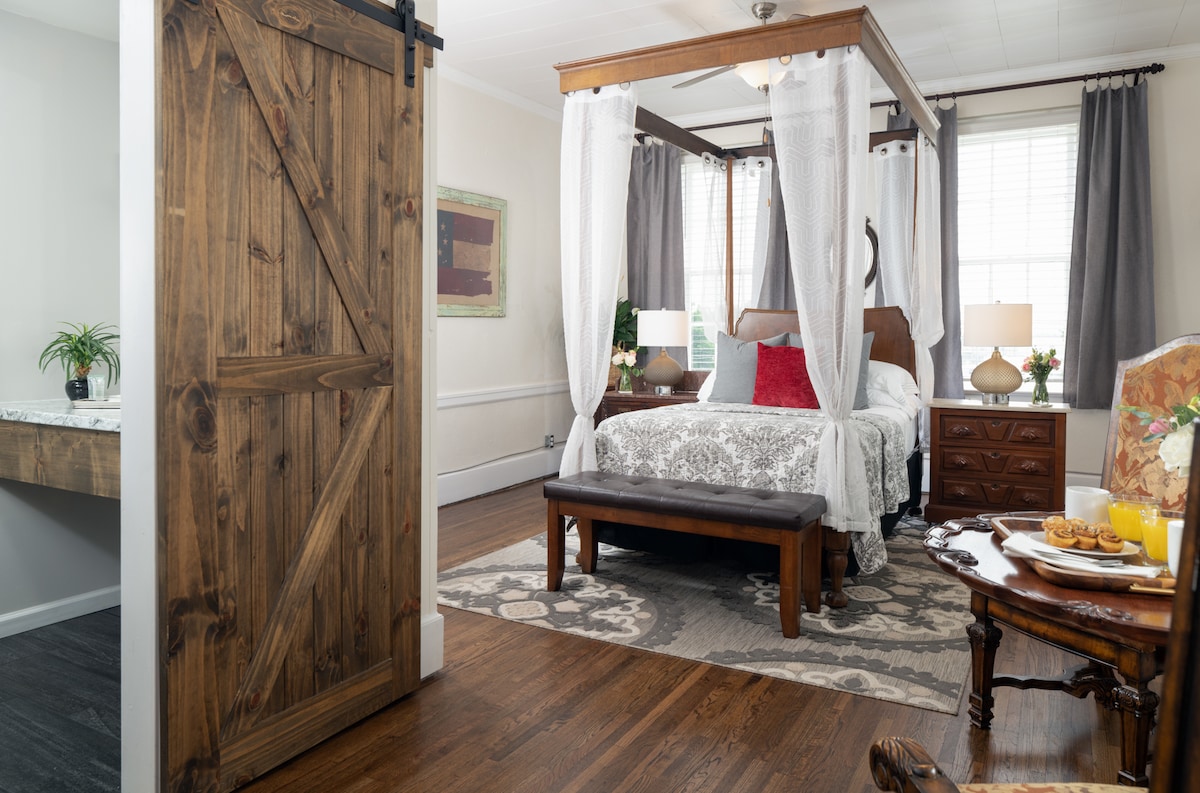 The room features a 4-poster bed draped with white fabric, complemented by bedside tables and lamps. A wooden bench sits at the foot of the bed, and a vintage chair enhances the decor. Natural light enters through the window, illuminating the hardwood floor and cozy surroundings.