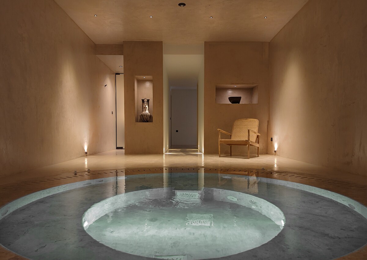 An indoor spa room features a heated plunge pool, surrounded by smooth concrete walls. Soft lighting enhances the serene atmosphere, while minimalist furniture is positioned nearby. Several niches house decorative items, contributing to a tranquil and relaxing environment.
