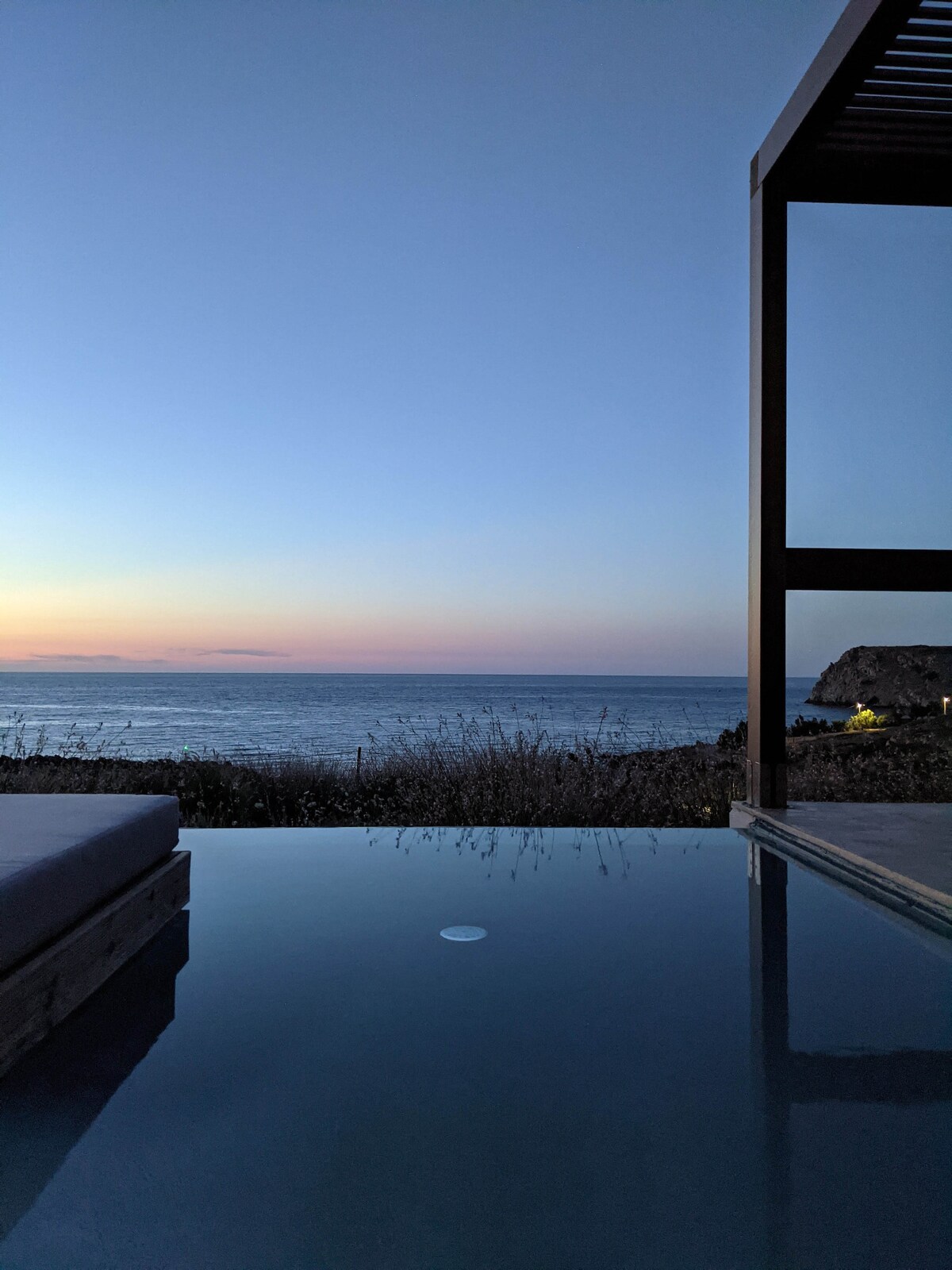 An infinity pool reflects the tranquil hues of a sunset over the sea, framed by a wooden pergola. The landscape transitions from land to water, creating a serene environment that invites relaxation.