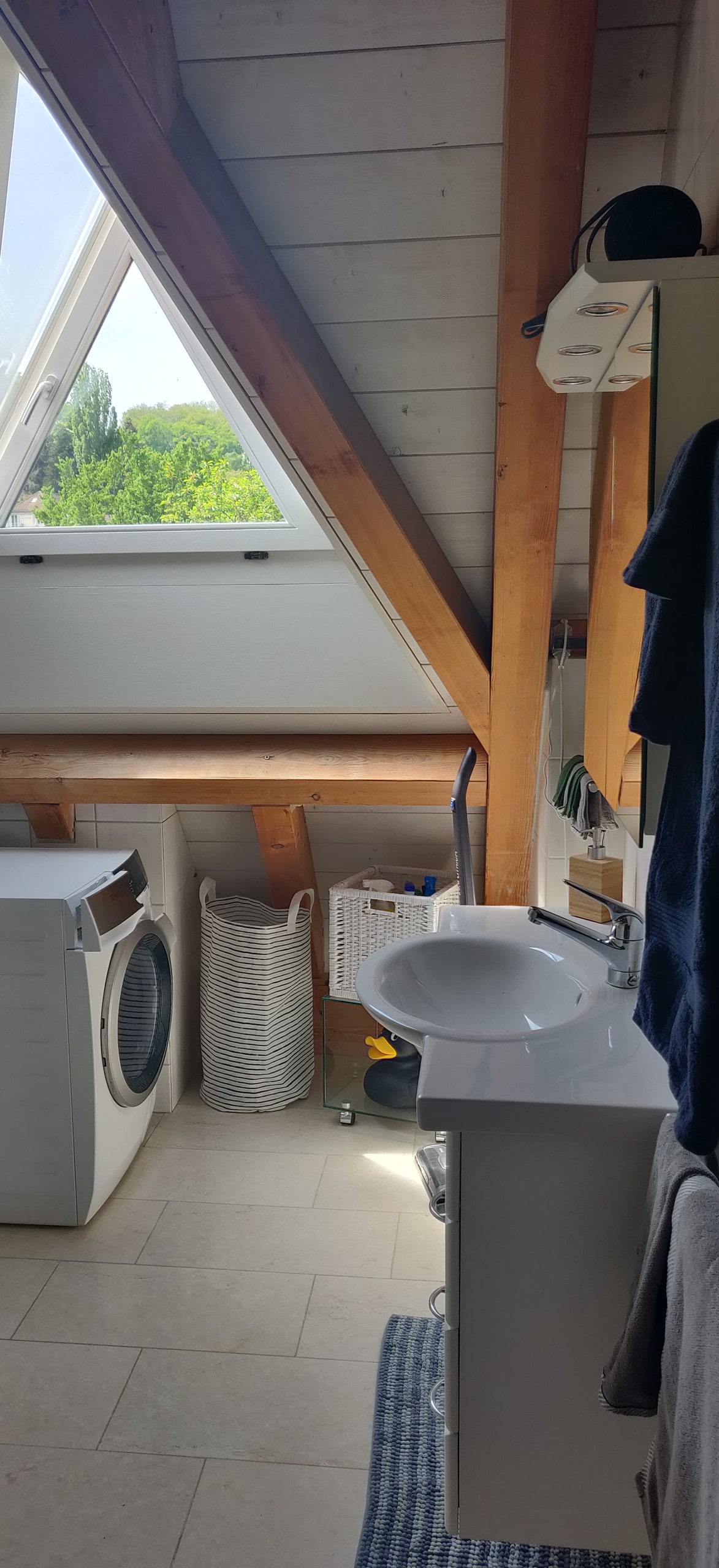A well-lit bathroom features a modern sink mounted on a white vanity, complemented by a washer and dryer. Natural light filters through a triangular window, highlighting wooden beams and storage baskets, creating an organized and functional space.