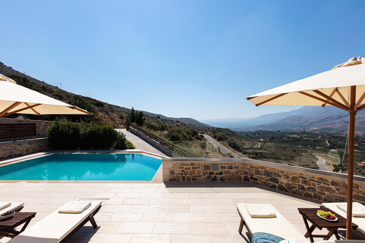 The image shows a private outdoor swimming pool surrounded by sun loungers under large umbrellas. A scenic view of the mountains and valley is presented in the background, with clear blue skies overhead, enhancing the serene atmosphere.