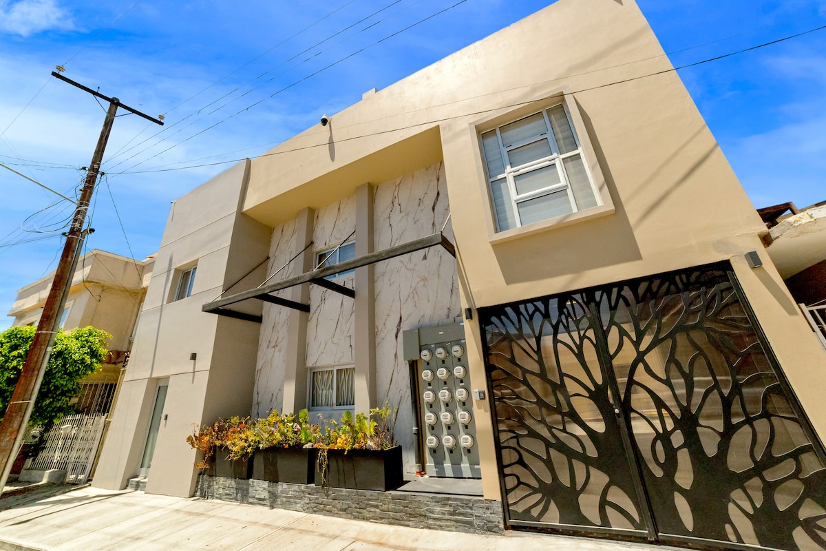 The modern building exterior features a sleek faƧade with a marble design and large windows. A decorative gate with a tree motif adds visual interest. Lush greenery is planted in front, enhancing the welcoming appearance of the entrance.