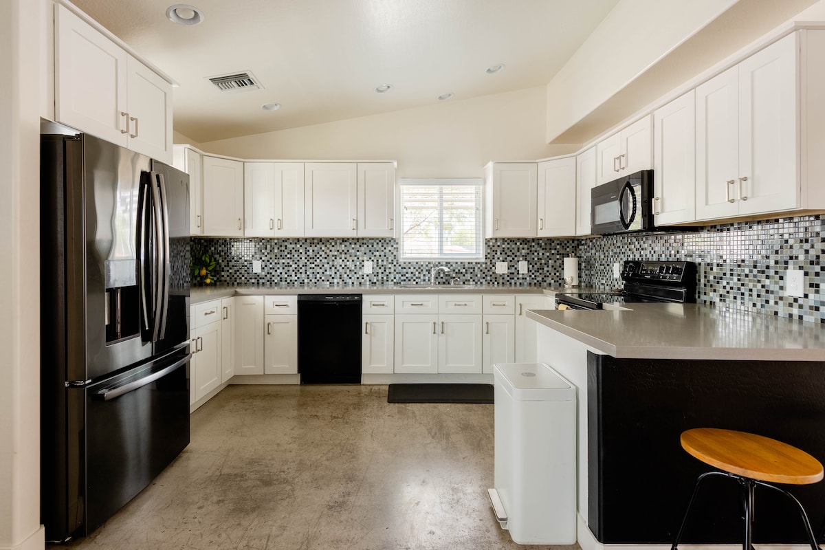The kitchen features white cabinetry with modern black appliances, including a refrigerator, oven, and microwave. A mosaic tile backsplash adds visual interest. A circular wooden stool is positioned next to a counter. Broad windows provide natural light, illuminating the spacious layout.