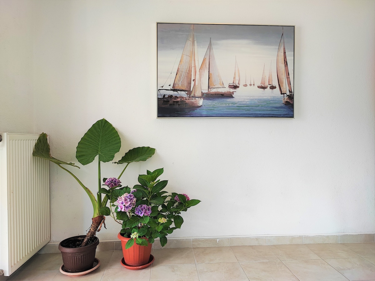A tranquil corner of the space features a large framed painting depicting sailing boats on calm waters. Potted plants, including a leafy green plant and blooming hydrangeas, add a touch of nature. A radiator is positioned against the white wall, enhancing the serene atmosphere.