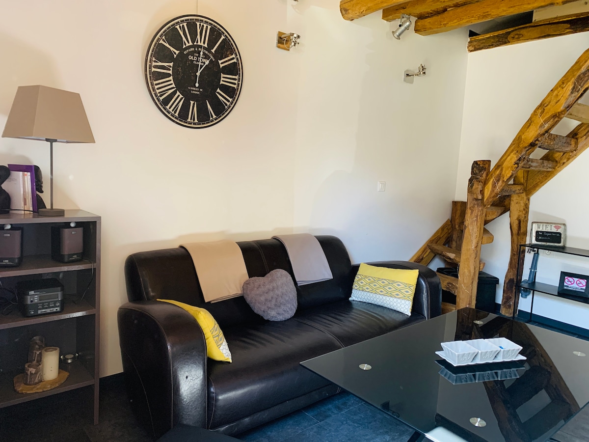 A cozy seating area features a dark leather sofa adorned with light-colored throw blankets and a decorative pillow. A glass table sits nearby, reflecting natural light, while wooden beams enhance the space's rustic charm. A large wall clock adds functionality and style.