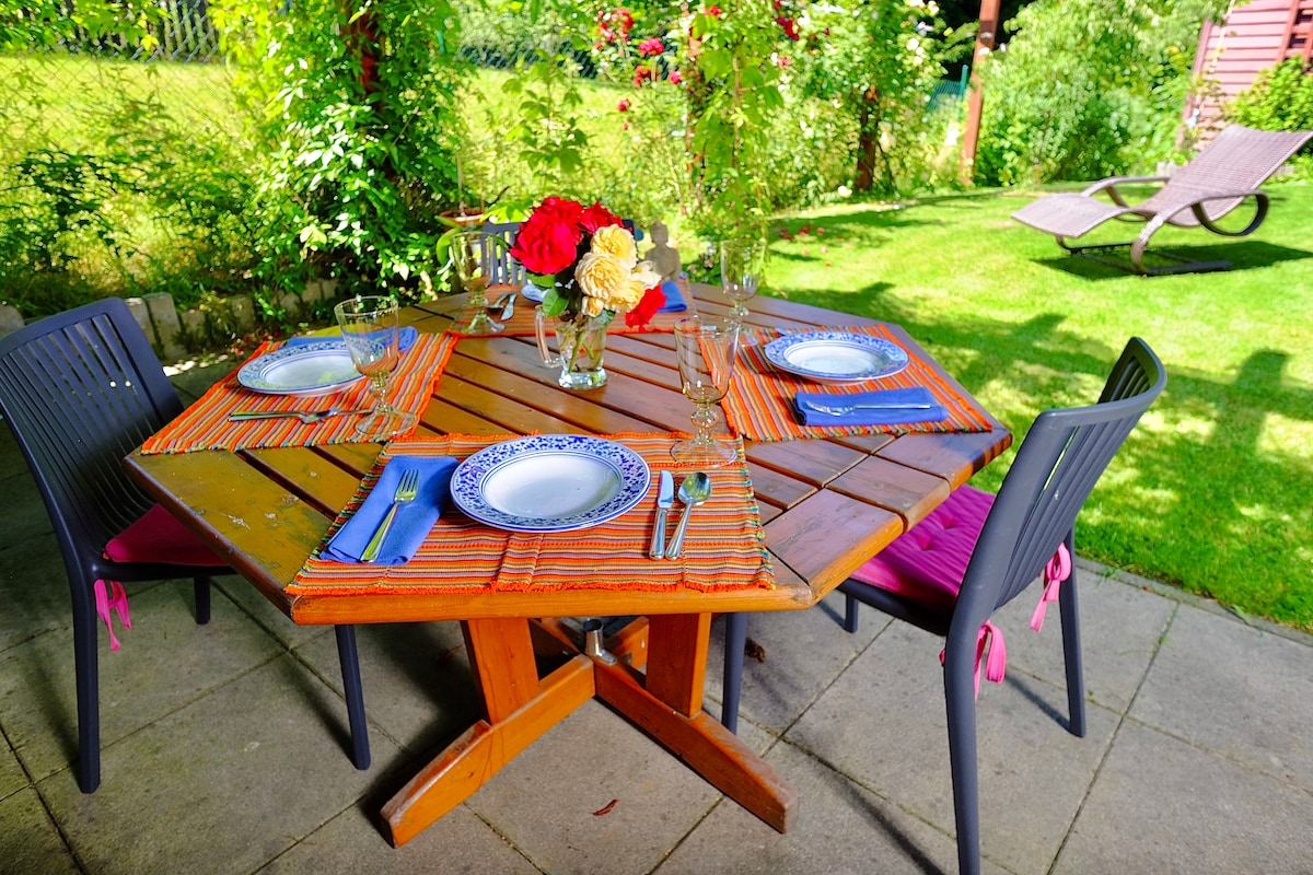 A charming outdoor dining area is presented, featuring a wooden table set with blue and white plates, cutlery, and colorful table runners. Flower arrangements add a vibrant touch. Surrounding greenery enhances the serene atmosphere, complemented by a lounging chair in the background.