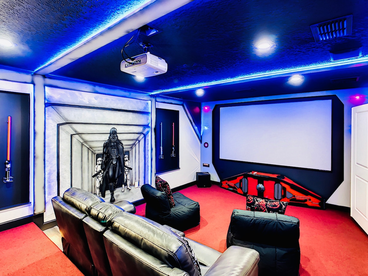 A themed movie theater features comfortable dark seating arranged in front of a large projection screen. The space is illuminated by blue LED lights, with character artwork adorning the walls, providing a unique cinematic environment.