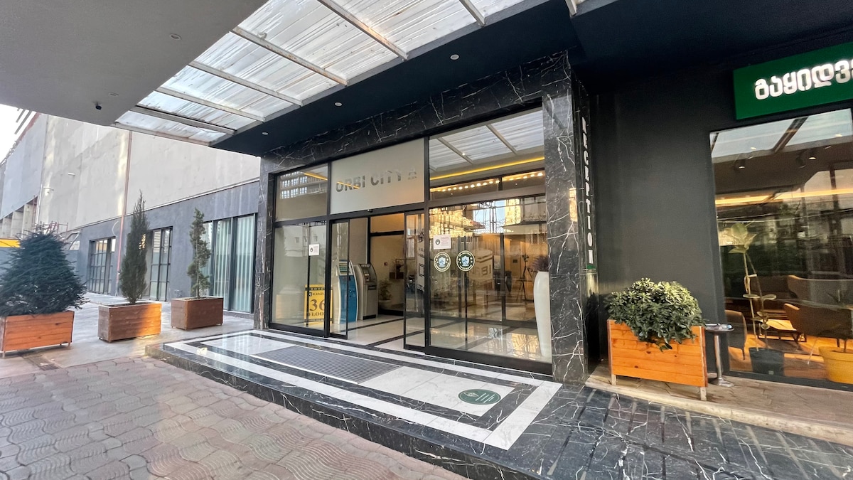The entrance of Orbi City Towers is displayed, featuring modern architecture with large glass doors. A tiled pathway leads to the entrance, while potted greenery is positioned on either side. The lobby area is visible through the glass, showcasing the welcoming atmosphere.