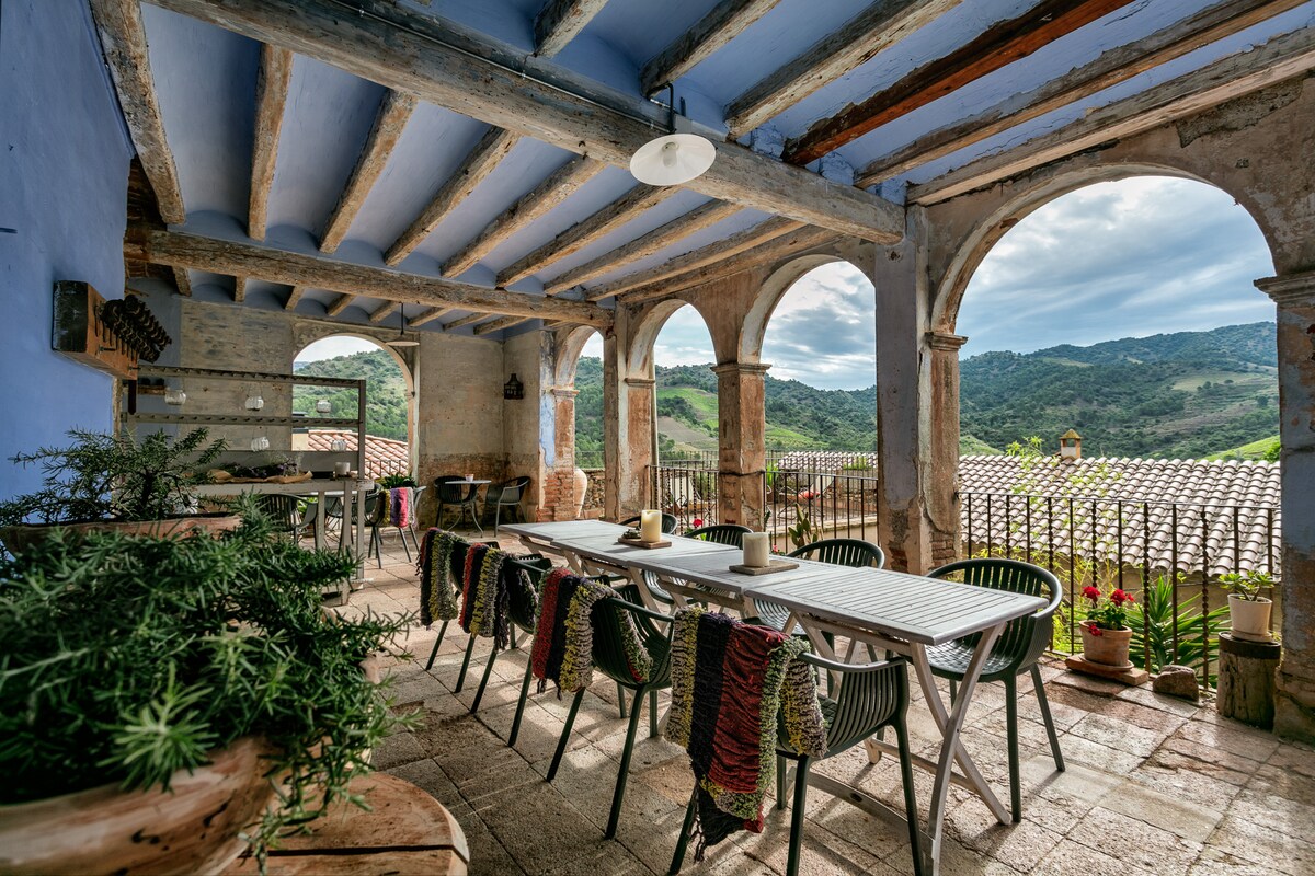 An outdoor terrace features multiple tables surrounded by stylish chairs and greenery. Arched stone columns frame the view of rolling hills and rooftops. The space is adorned with rustic decor, highlighting the inviting atmosphere for gatherings and enjoying nature.