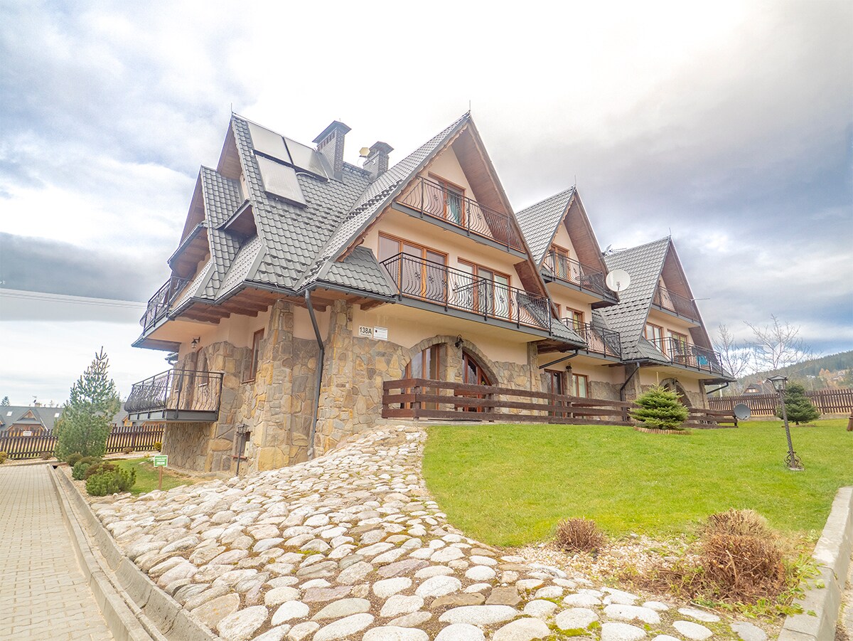 The exterior of a three-story building features a stone facade and wooden balconies, surrounded by landscaped grounds. A paved pathway leads to the entrance, flanked by small trees and shrubs. The structure showcases a traditional architectural style typical of mountain lodges.