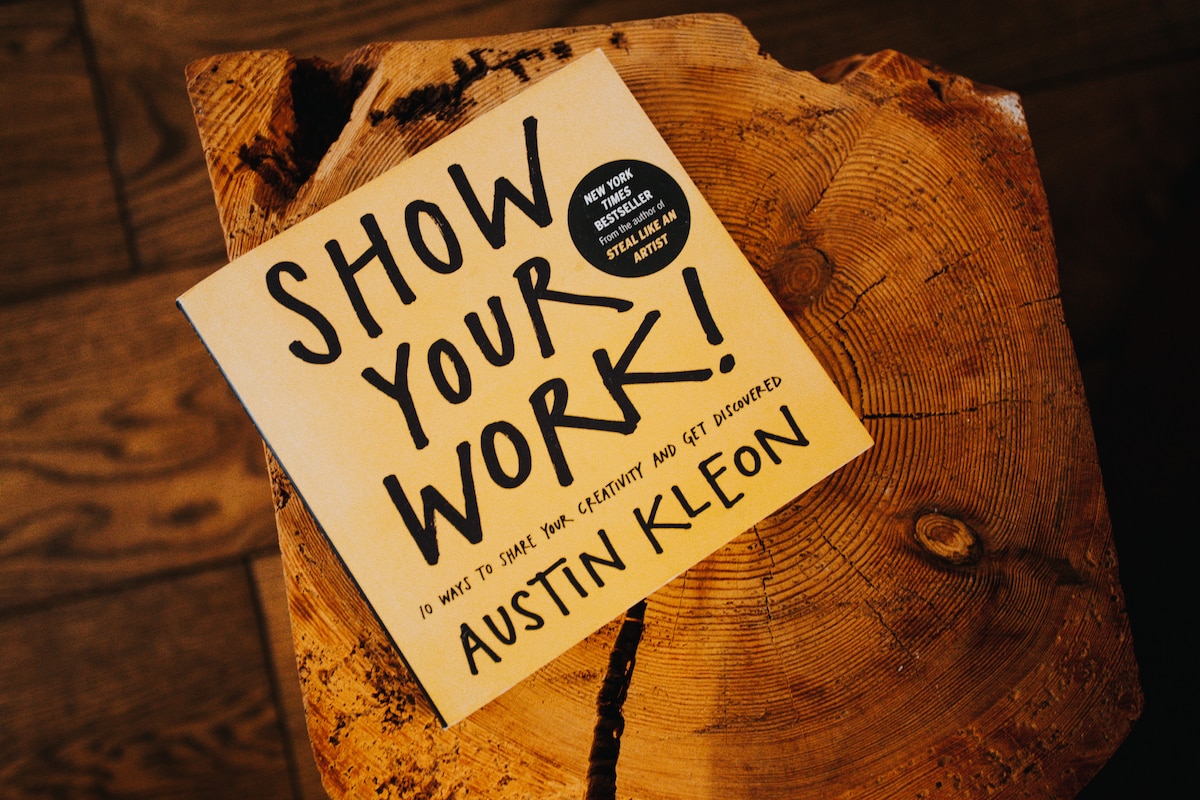 A book titled 'Show Your Work!' by Austin Kleon is placed on a rustic wooden surface. The cover features bold black text on a light background, emphasizing its title. The natural texture of the wood adds warmth to the setting.