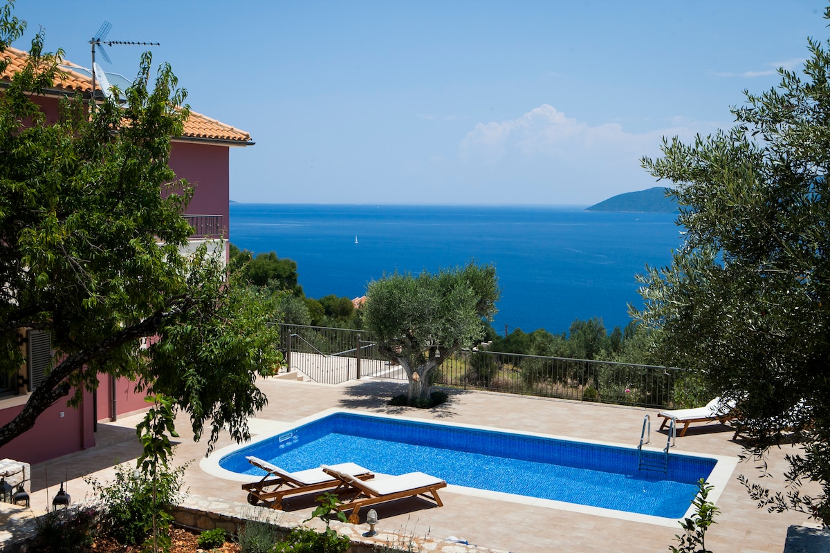 A clear view of a private swimming pool framed by landscaped gardens, featuring two loungers positioned beside the water. In the background, the expansive sea reflects the blue sky, complemented by lush greenery and mature trees, offering a serene and secluded atmosphere.