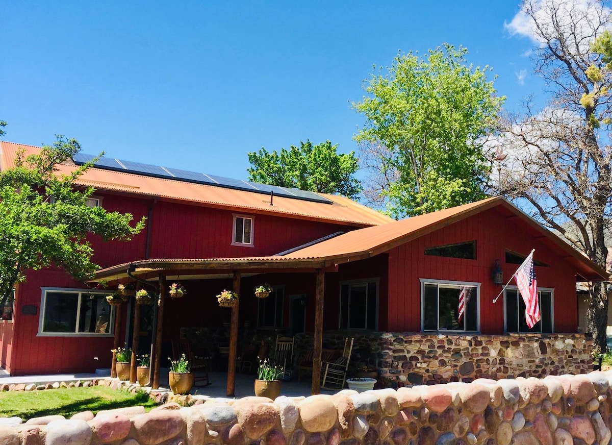 The exterior of the property features a red facade with a rustic stone base. A covered porch with hanging flower pots provides a welcoming entrance, while solar panels are visible on the roof. Two American flags flutter in the gentle breeze beneath a bright blue sky.