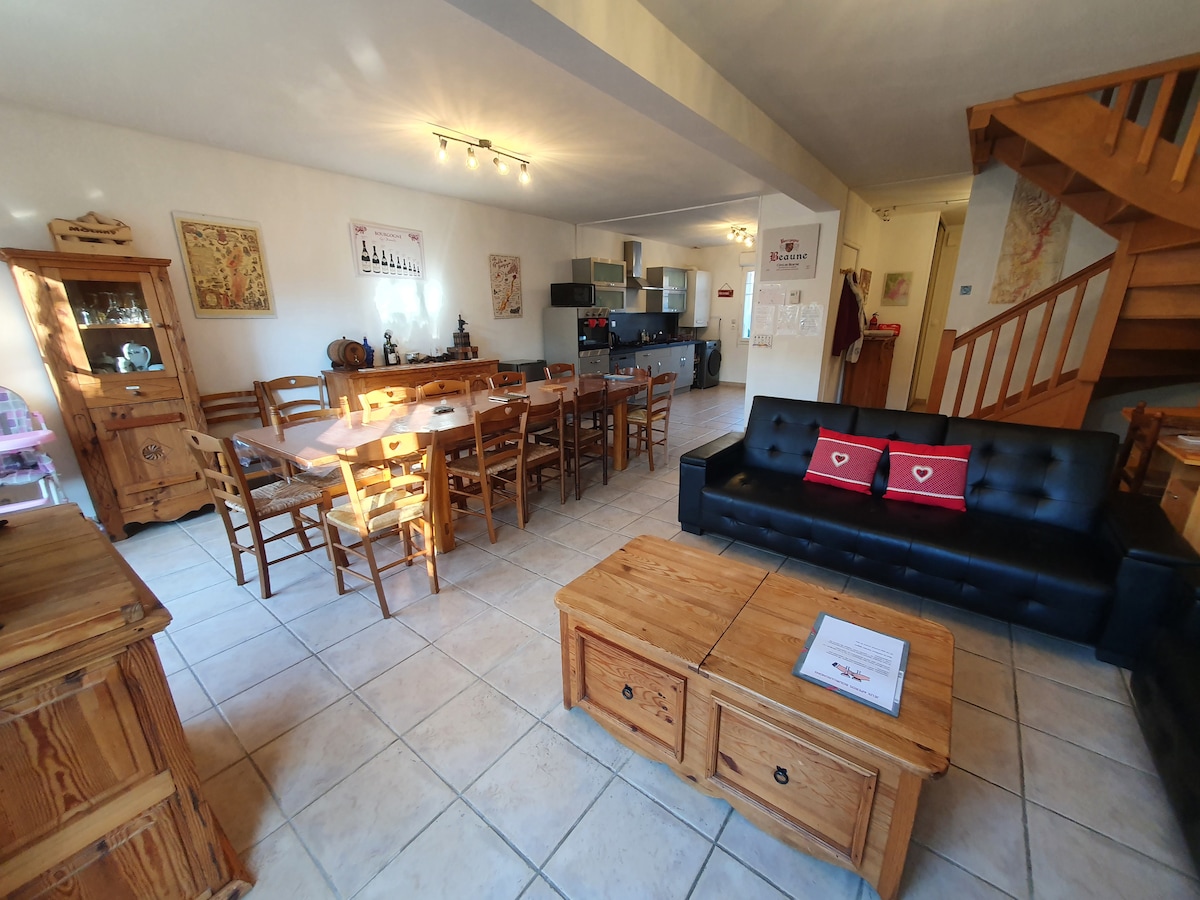 A spacious living area is presented featuring a wooden dining table surrounded by chairs. A black sofa with red cushions sits opposite a wooden coffee table. The open kitchen area is visible, equipped with modern appliances and ample counter space. Natural light illuminates the room through multiple windows.