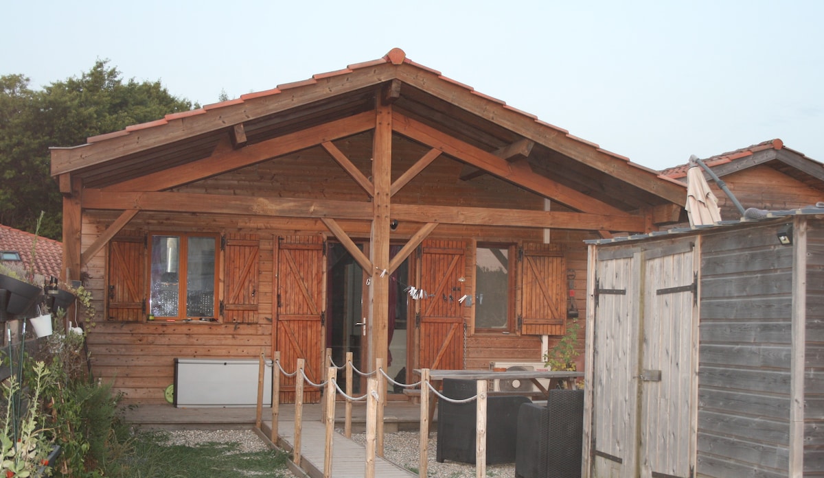 A wooden house features an expansive porch area with a railing leading to the entrance. Shuttered windows provide ample light, while a nearby wooden shed offers additional storage. The natural wood exterior complements the surrounding greenery, creating a harmonious outdoor space.