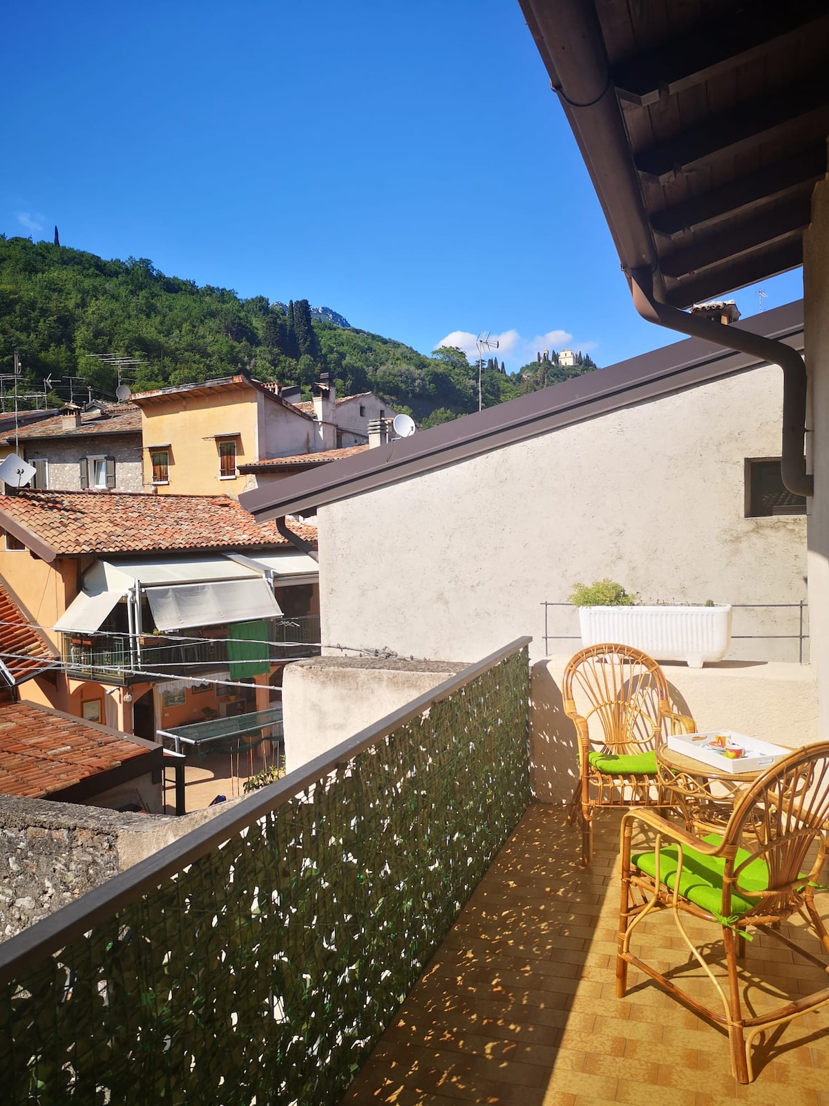 A balcony offers views of neighboring rooftops and greenery, surrounded by a mountain landscape. Two wicker chairs and a small table are positioned on the space, inviting relaxation under clear blue skies.