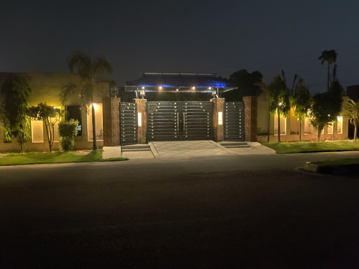 The entrance gate is illuminated by soft lights, emphasizing the modern design and featuring a sleek, dark facade. Lush greenery is visible on either side, enhancing the welcoming aspect of the property at night.