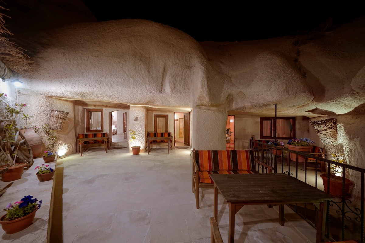 A spacious outdoor area is illuminated softly, showcasing a blend of natural cave formations and comfortable seating arrangements. Potted plants with vibrant flowers add a touch of color, while sturdy wooden tables and chairs offer a relaxed spot to enjoy the surroundings.