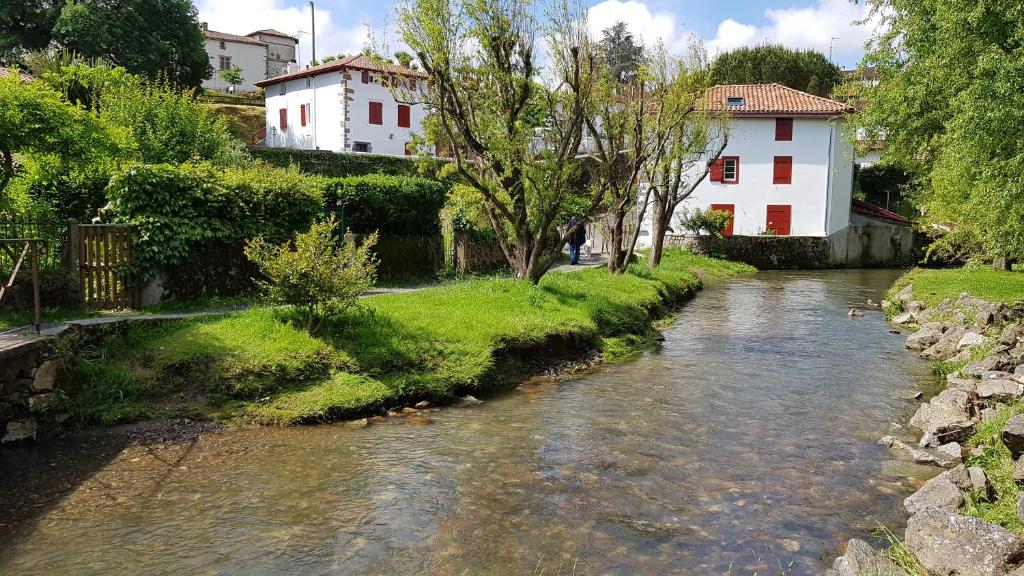 A serene landscape is depicted featuring a gently flowing river bordered by lush greenery. A white building with red windows is seen in the background, complementing the vibrant natural surroundings and creating a tranquil atmosphere.