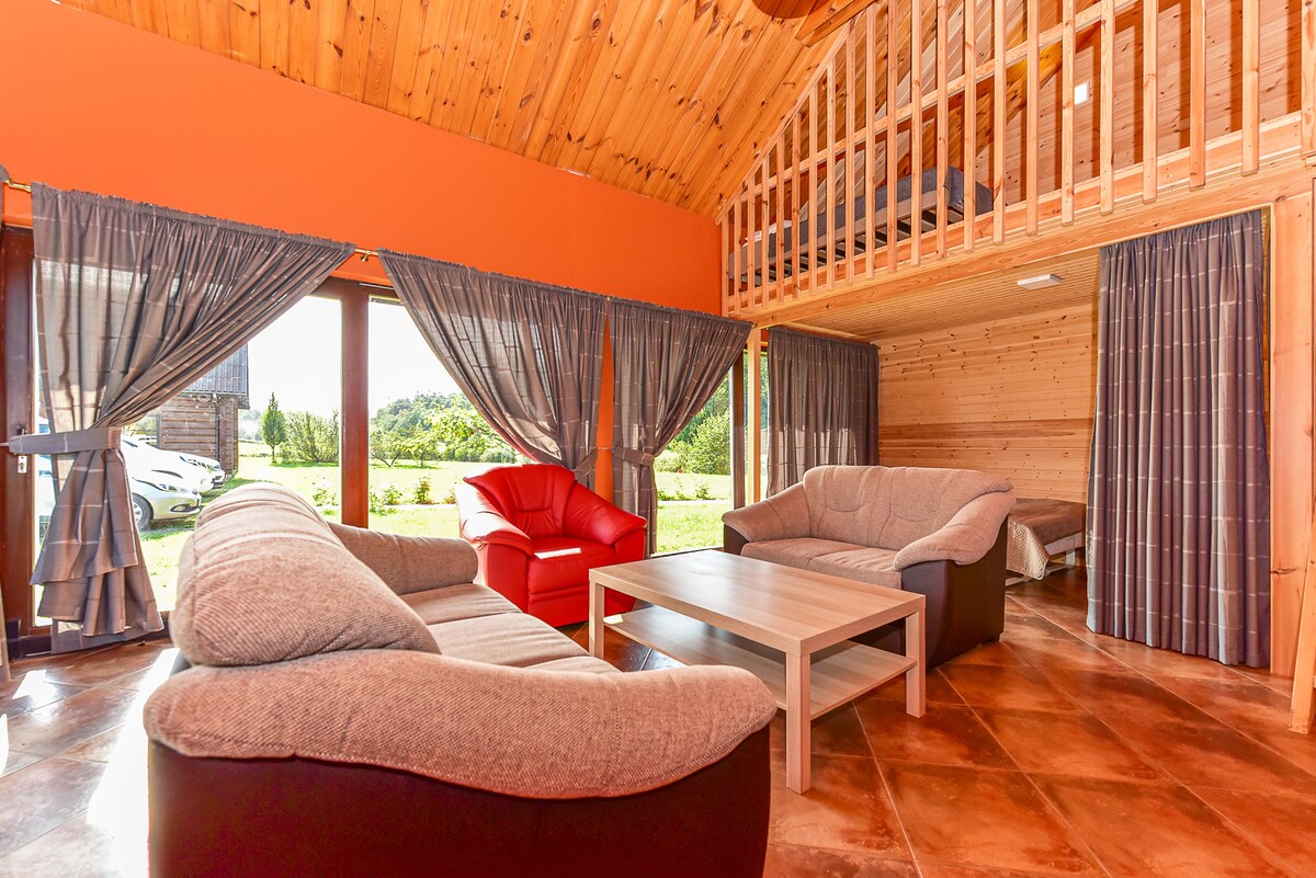A spacious living area is highlighted by large windows, allowing natural light to fill the space. Comfortable seating arrangements include two sofas and an armchair. A wooden loft can be seen above, adding an open feel to the room, with greenery visible outside.