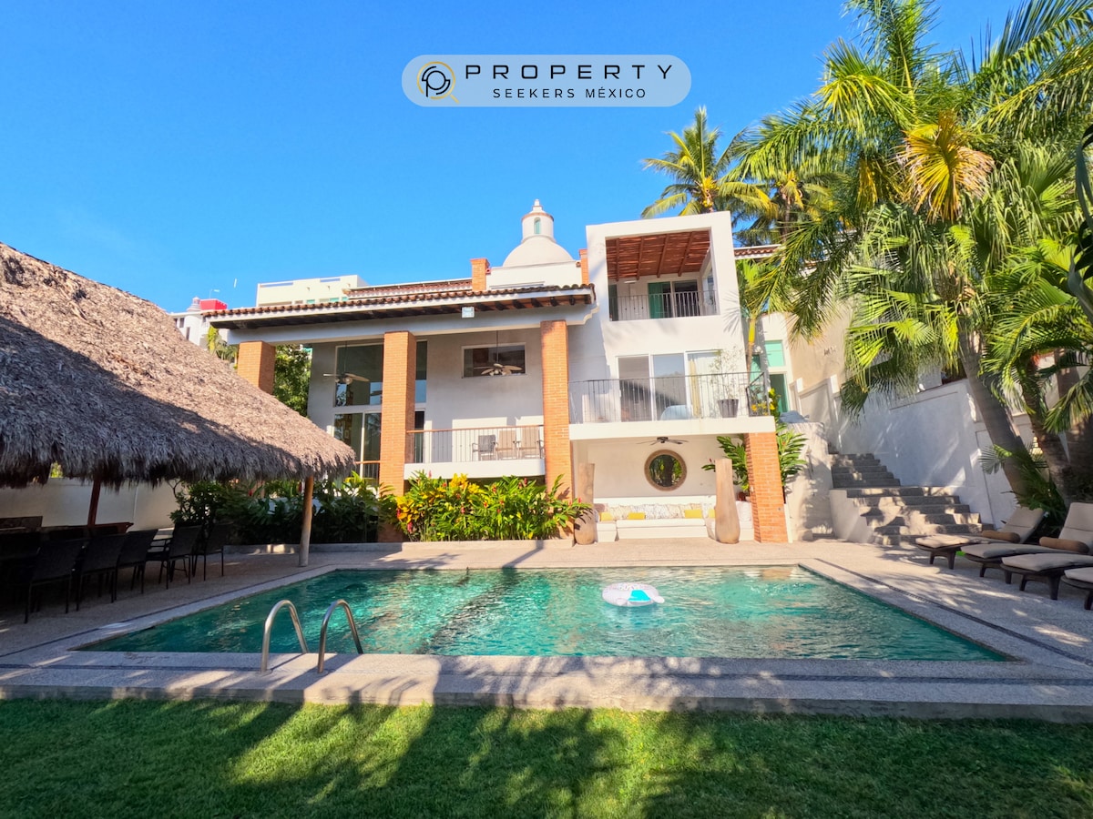 The villa is presented with a lush garden and a sparkling pool, framed by palm trees. A spacious terrace, nestled beneath thatched roofing, provides seating. The two-story structure showcases large windows and a balcony, enhancing the inviting outdoor space for relaxation.
