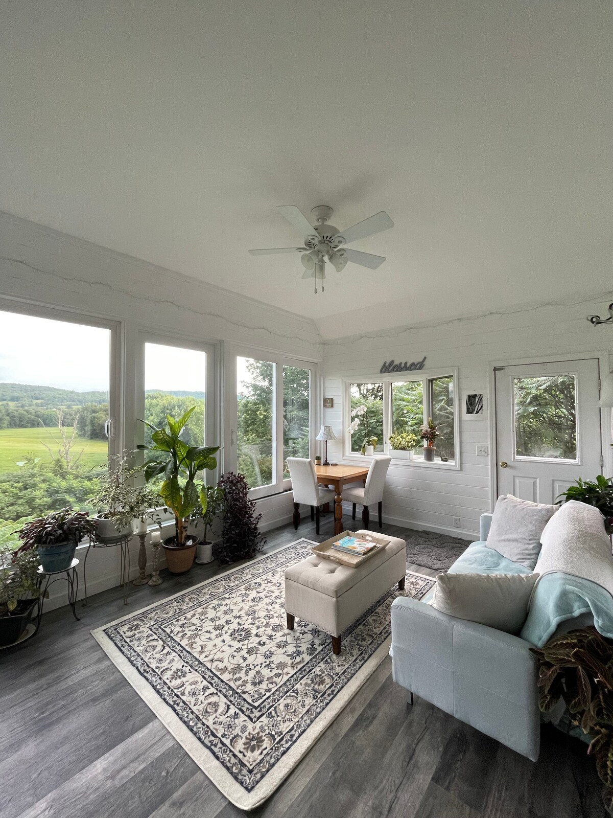 A sunroom filled with natural light features large windows offering views of the surrounding greenery. A small dining table with two chairs sits alongside a comfortable seating area, accented by a decorative rug and potted plants, creating a serene atmosphere.