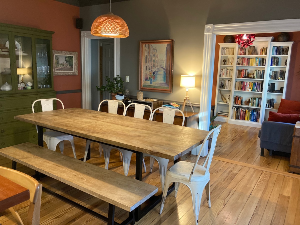 A spacious dining area features a wooden table with a rustic finish and white metal chairs. A large green cabinet stands against the wall, complemented by a warm pendant light overhead. In the background, a cozy living area with bookshelves filled with books is visible.