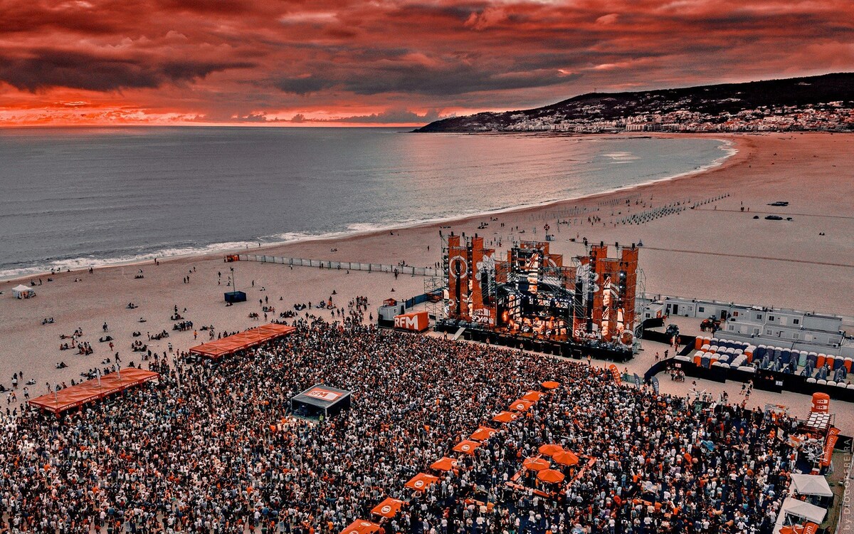 A large crowd gathers on the beach, with a stage set up in the foreground. The calm sea reflects a dramatic sky, characterized by vivid red and orange hues. People can be seen enjoying themselves along the shoreline and on the beach.