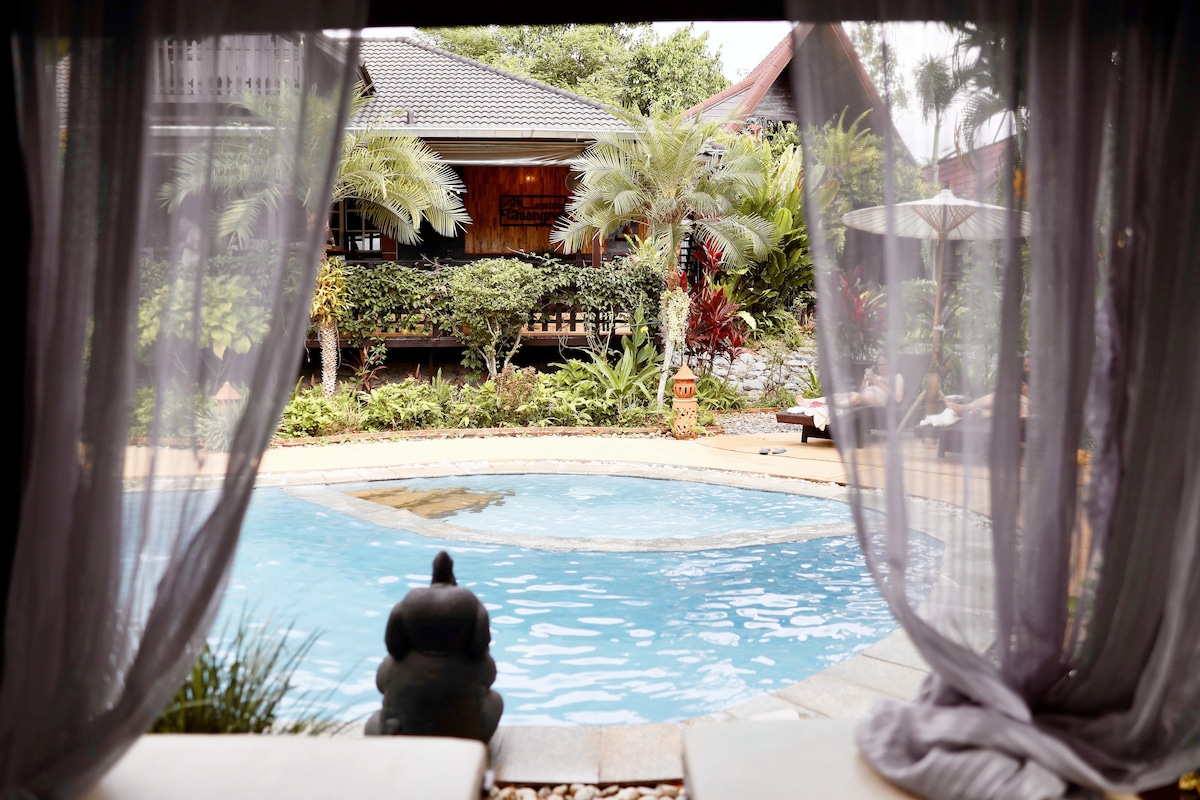 A serene view is framed by sheer curtains, revealing a tranquil swimming pool surrounded by lush greenery. Decorative elements such as a stone figure add a touch of charm. The backdrop features a wooden structure with landscaped gardens for a peaceful ambiance.