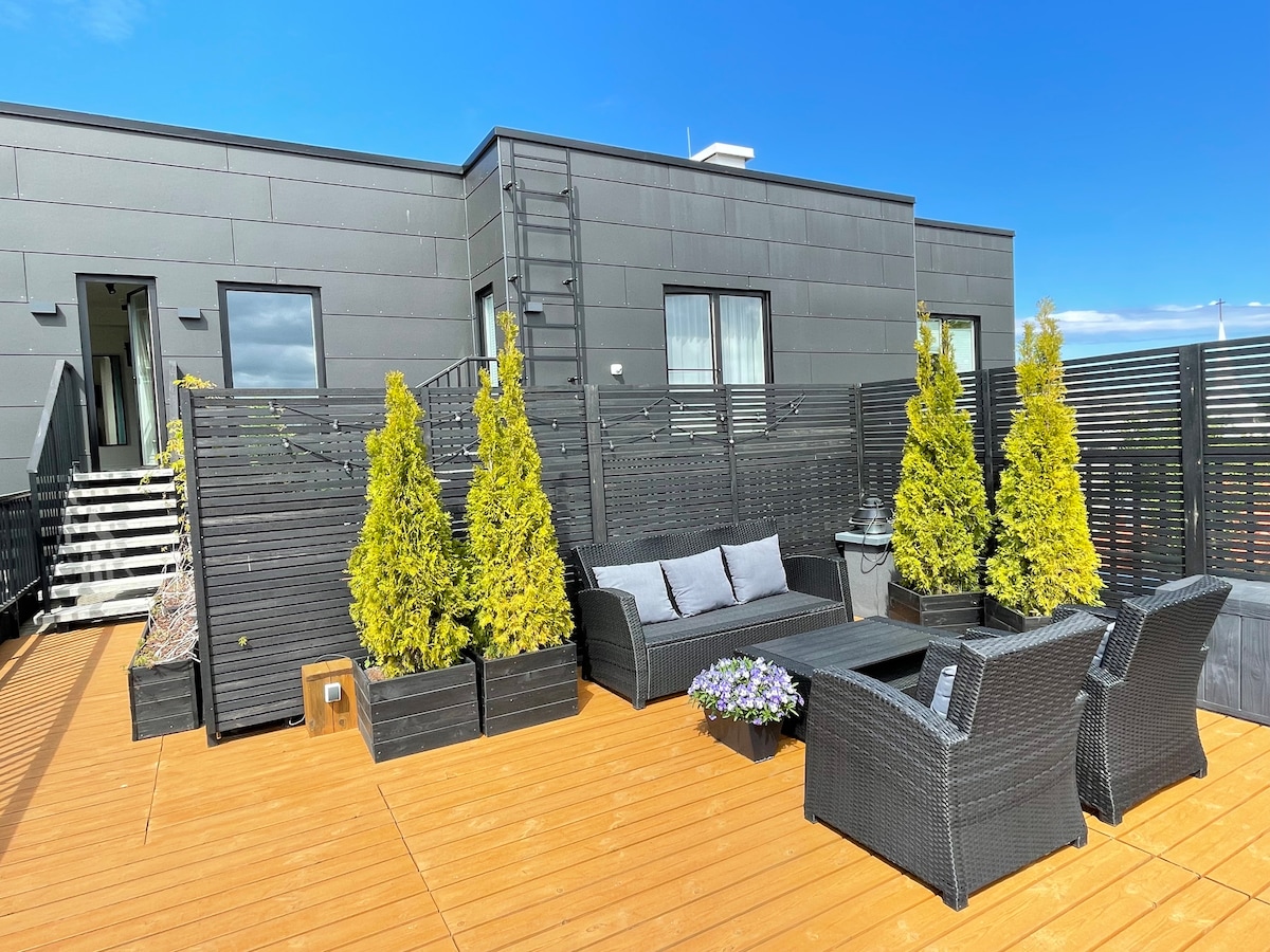 A spacious private terrace showcases wooden decking complemented by modern outdoor furniture, including a seating area and coffee table. Potted shrubs add greenery, enhancing the space's appeal. The entrance to the apartment is visible, along with a staircase leading to the terrace.