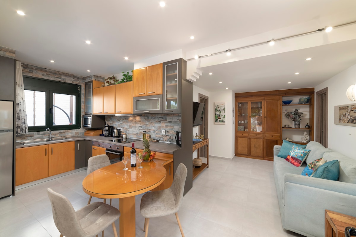 A modern kitchen area is featured, with light wooden cabinetry and stone accents. A round dining table with soft grey chairs is present, set for a meal. The living space shows a light blue sofa and decorative items on shelves, contributing to a welcoming atmosphere.