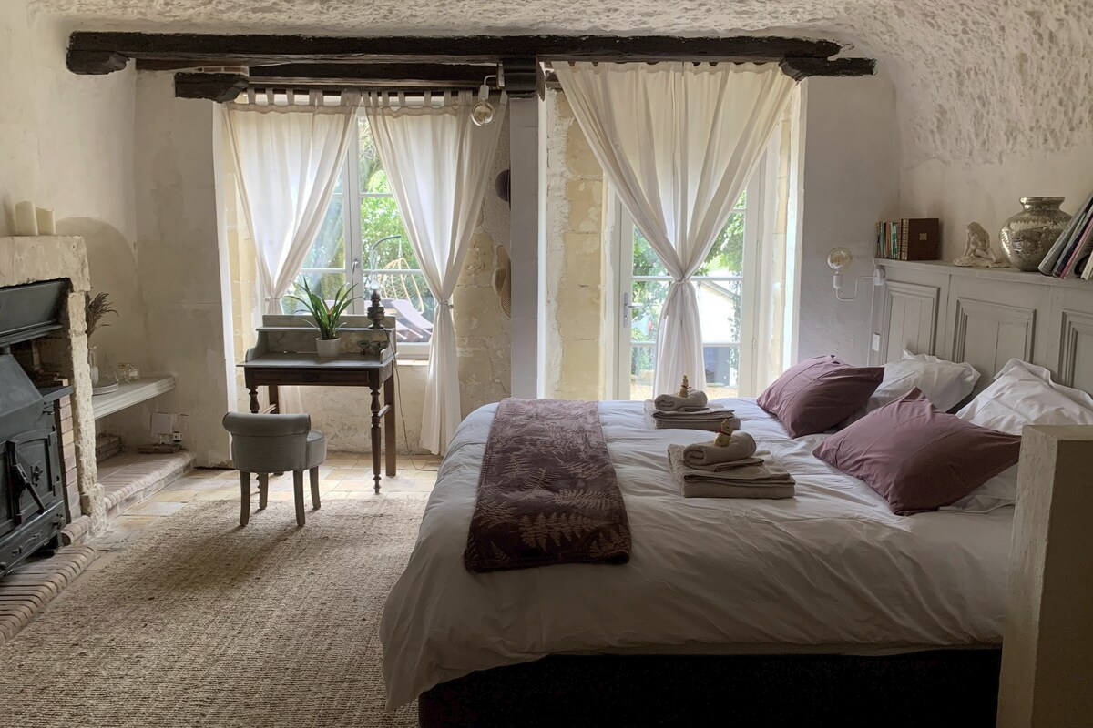 A serene bedroom is showcased with a spacious bed dressed in neutral linens, flanked by light-filled windows draped with sheer curtains. A cozy seating area includes a small chair and decorative table. A fireplace adds a rustic element to the warm interior.