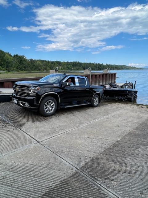 A black pickup truck is seen at the boat launch area, with a boat trailer attached, positioned on a concrete ramp leading to the calm waters of Lake Champlain. Lush greenery and a clear blue sky with scattered clouds create a serene backdrop.