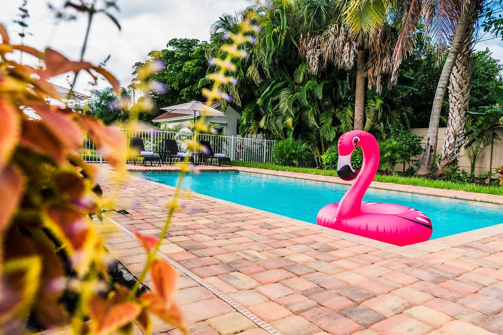 A vibrant pink flamingo float is situated in a sparkling blue pool, surrounded by lush greenery and palm trees. The outdoor space is framed by a well-maintained patio, featuring a relaxing atmosphere under partly cloudy skies.