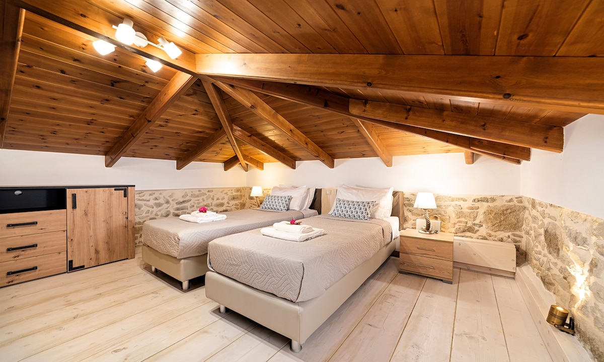 A spacious bedroom features two twin beds with light bedding arranged for comfort. Wooden beams stretch across the ceiling, paired with soft lighting from bedside lamps. A stone accent wall adds a rustic touch, while minimal furnishings create an inviting atmosphere.