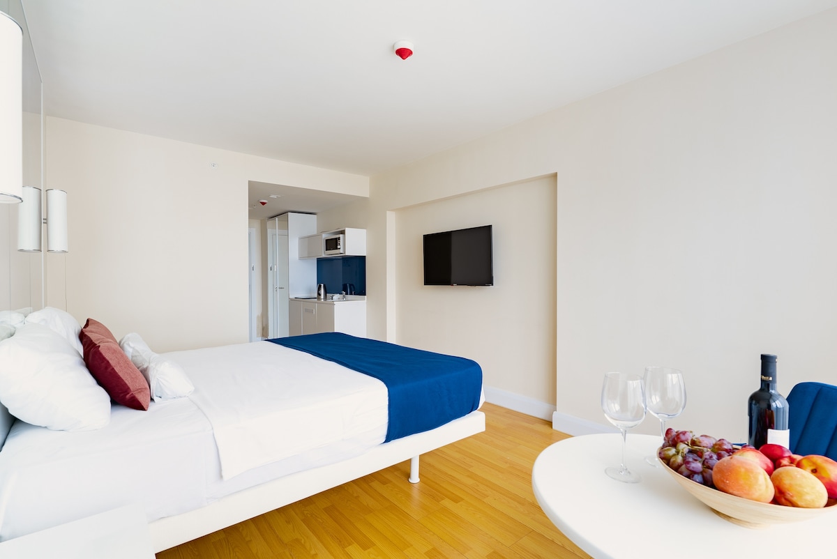 A spacious studio apartment is presented, featuring a comfortable bed with white linens and accent pillows in red. A compact dining area with a round table is set with a bottle of wine and a variety of fruits. A kitchenette and television are visible, enhancing the functionality.